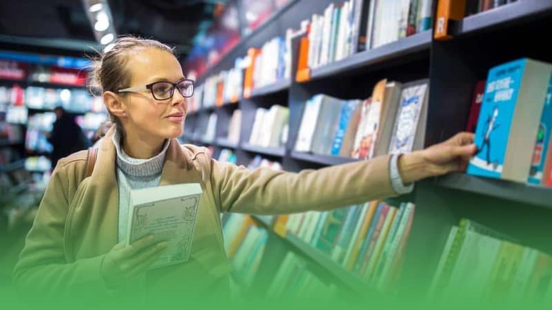 Female bookstore owner in glasses peruses books on display.