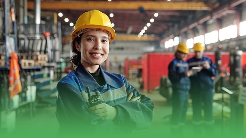 Woman in hard hat and blue overalls stands in front of factory at manufacturing premises.