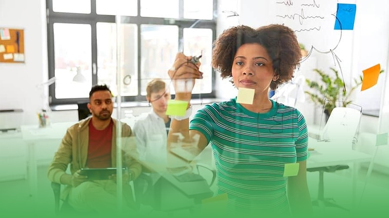 Woman writes on whiteboard in startup business surrounded by other people in the background.