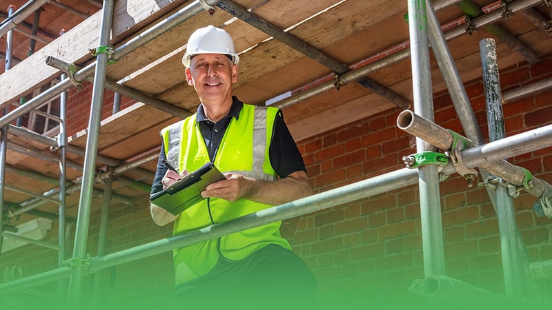 Construction business owner wearing hard hat and safety vest stands on scaffolding at worksite.