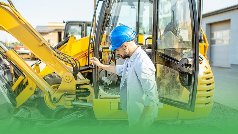 Man in hard hat and blue shirt examines yellow excavator representing excavation business owner.