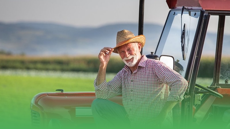 Farm equipment business owner sits on tractor in field wearing hat.