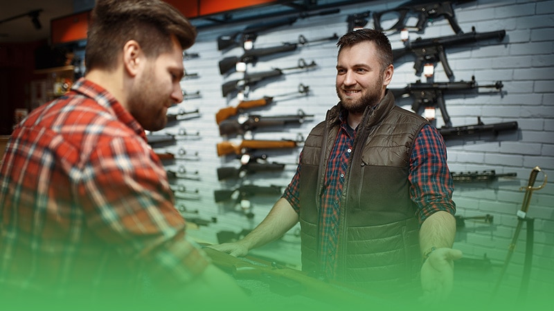 Gun store owner and customer examining firearms in store.