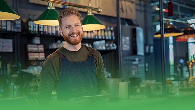 Restaurant business owner wearing green apron smiles while confidently holding tablet.